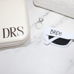 K&ouml;nnte beinhalten: Eine wei&szlig;e Kosmetiktasche mit den schwarzen Initialen "DRS", ein silberner Verlobungsring und eine wei&szlig;e Sonnenbrille mit dem Wort "BRIDE" darauf. Die Artikel liegen auf einer wei&szlig;en Marmoroberfl&auml;che.