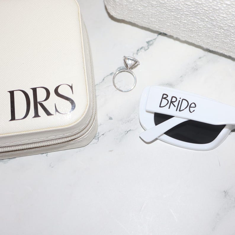 K&ouml;nnte beinhalten: Eine wei&szlig;e Kosmetiktasche mit den schwarzen Initialen "DRS", ein silberner Verlobungsring und eine wei&szlig;e Sonnenbrille mit dem Wort "BRIDE" darauf. Die Artikel liegen auf einer wei&szlig;en Marmoroberfl&auml;che.