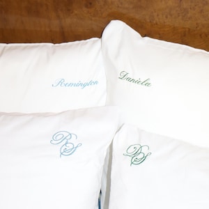 Puede incluir: Cuatro almohadas blancas con texto e iniciales bordadas. El texto "Daniela" est&aacute; en escritura verde, "Remington" en escritura azul claro, y las iniciales "B S" y "D S" en azul claro y verde.
