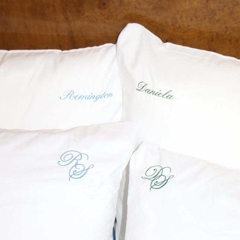 Puede incluir: Cuatro almohadas blancas con texto e iniciales bordadas. El texto "Daniela" est&aacute; en escritura verde, "Remington" en escritura azul claro, y las iniciales "B S" y "D S" en azul claro y verde.