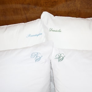 Puede incluir: Cuatro almohadas blancas con detalles bordados. Dos almohadas tienen nombres bordados con hilo azul y verde. Las otras dos almohadas tienen iniciales bordadas con hilo azul y verde. Las almohadas est&aacute;n sobre una superficie de madera.