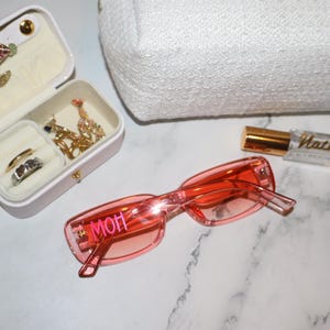 K&ouml;nnte beinhalten: Rosafarbene Sonnenbrille mit dem Wort "MOH" auf dem Rahmen. Eine kleine Schmuckschatulle mit Ringen und Halsketten ist zu sehen, ebenso wie eine wei&szlig;e, strukturierte Kosmetiktasche und eine Parf&uuml;mflasche.
