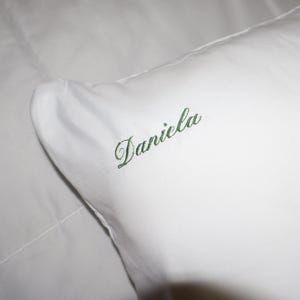 Puede incluir: Una almohada blanca con el nombre "Daniela" bordado en verde. El nombre est&aacute; escrito en cursiva. La almohada est&aacute; hecha de una tela suave y lisa, lo que sugiere un conjunto de ropa de cama personalizado.