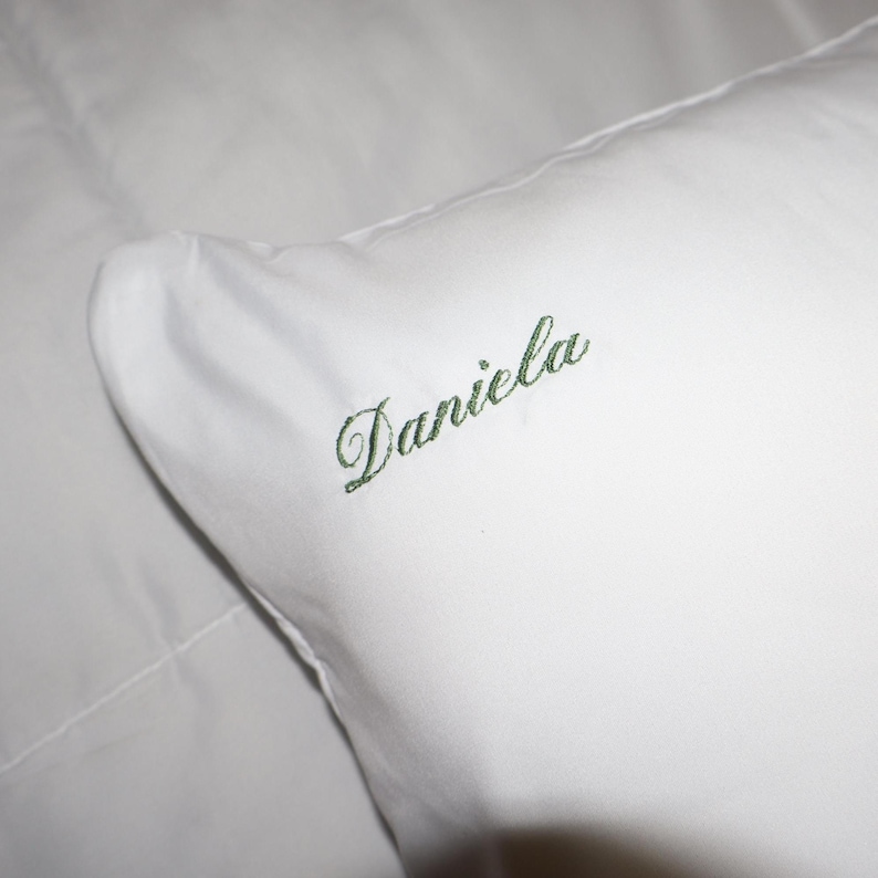 Puede incluir: Una almohada blanca con el nombre "Daniela" bordado en verde. El nombre est&aacute; escrito en cursiva. La almohada est&aacute; hecha de una tela suave y lisa, lo que sugiere un conjunto de ropa de cama personalizado.