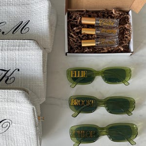 K&ouml;nnte beinhalten: Eine Auswahl an personalisierten Geschenken, darunter wei&szlig;e, strukturierte Kosmetiktaschen mit schwarzen, gestickten Initialen, gr&uuml;ne Sonnenbrillen mit goldfarbenen Buchstaben und eine Schachtel mit Parf&uuml;mflaschen. Die Sonnenbrillen sind mit "Ellie", "Brooke" und "Chloe" beschriftet.