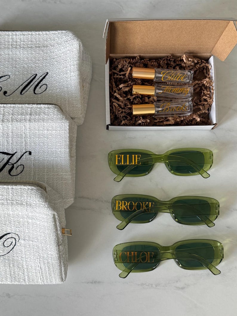 K&ouml;nnte beinhalten: Eine Auswahl an personalisierten Geschenken, darunter wei&szlig;e, strukturierte Kosmetiktaschen mit schwarzen, gestickten Initialen, gr&uuml;ne Sonnenbrillen mit goldfarbenen Buchstaben und eine Schachtel mit Parf&uuml;mflaschen. Die Sonnenbrillen sind mit "Ellie", "Brooke" und "Chloe" beschriftet.