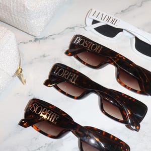 K&ouml;nnte beinhalten: Sonnenbrillen mit personalisierten Namen. Drei Sonnenbrillen im Schildpatt-Look mit den Namen "Boston", "Loren" und "Sophie" in goldfarbenen Buchstaben. Eine wei&szlig;e Sonnenbrille mit dem Namen "Maddie" in goldfarbenen Buchstaben. Eine wei&szlig;e Tasche im Hintergrund.