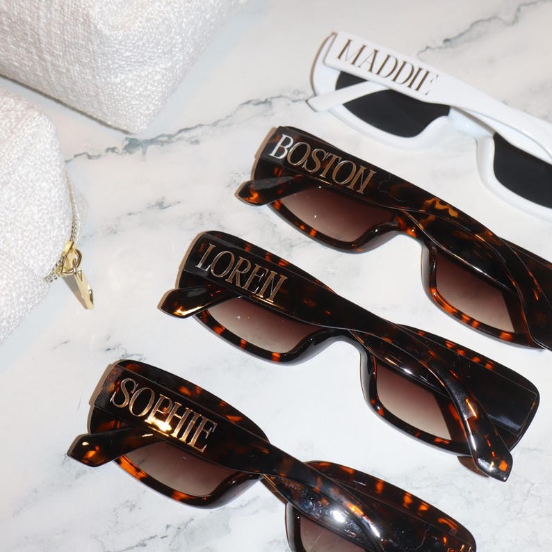 K&ouml;nnte beinhalten: Sonnenbrillen mit personalisierten Namen. Drei Sonnenbrillen im Schildpatt-Look mit den Namen "Boston", "Loren" und "Sophie" in goldfarbenen Buchstaben. Eine wei&szlig;e Sonnenbrille mit dem Namen "Maddie" in goldfarbenen Buchstaben. Eine wei&szlig;e Tasche im Hintergrund.