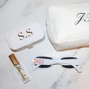 K&ouml;nnte beinhalten: Eine Sammlung wei&szlig;er Accessoires, darunter ein kleiner glitzernder Koffer mit goldenen Buchstaben "S.S", eine wei&szlig;e Kosmetiktasche mit gestickten Initialen und eine wei&szlig;e Sonnenbrille mit "BRIDE" in Rot. Eine kleine Parf&uuml;mflasche ist ebenfalls vorhanden.