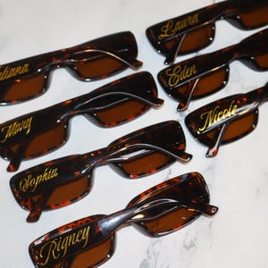 K&ouml;nnte beinhalten: Sieben Sonnenbrillen mit Schildpattmuster und braunen Gl&auml;sern sind auf einer wei&szlig;en Oberfl&auml;che angeordnet. Jede Brille hat einen anderen Namen in goldener Schrift auf dem oberen Rahmen: Katiana, Mary, Sophia, Laura, Eden, Nicole und Rigney.