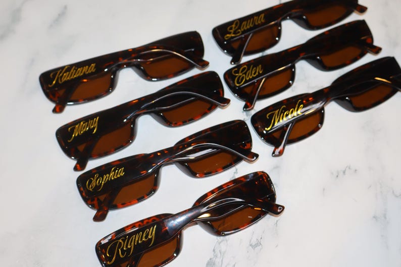K&ouml;nnte beinhalten: Sieben Sonnenbrillen mit Schildpattmuster und braunen Gl&auml;sern sind auf einer wei&szlig;en Oberfl&auml;che angeordnet. Jede Brille hat einen anderen Namen in goldener Schrift auf dem oberen Rahmen: Katiana, Mary, Sophia, Laura, Eden, Nicole und Rigney.