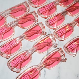 K&ouml;nnte beinhalten: Eine Ansammlung von rosafarbenen Sonnenbrillen mit transparenten Rahmen. Jede Brille hat einen anderen Namen, der in Rosa &uuml;ber dem Rahmen aufgedruckt ist. Die Sonnenbrillen sind auf einer wei&szlig;en Oberfl&auml;che angeordnet.