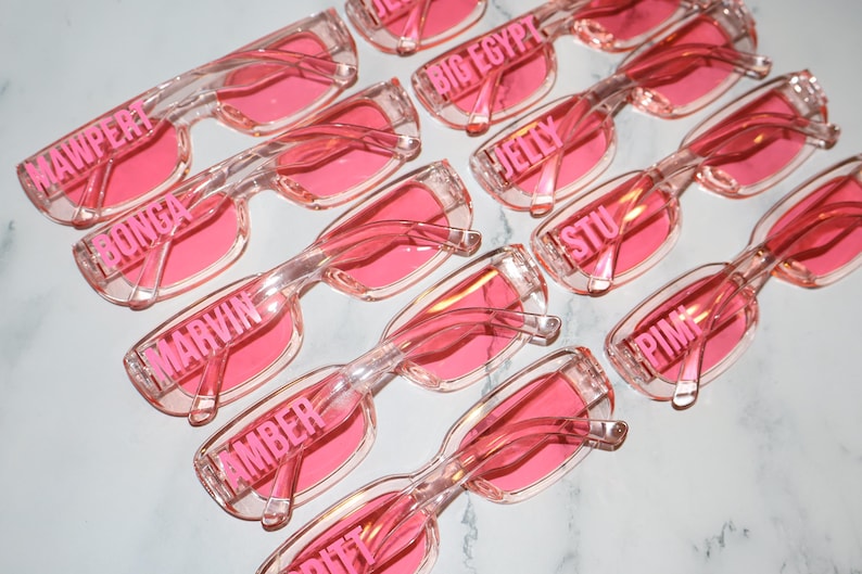 K&ouml;nnte beinhalten: Eine Ansammlung von rosafarbenen Sonnenbrillen mit transparenten Rahmen. Jede Brille hat einen anderen Namen, der in Rosa &uuml;ber dem Rahmen aufgedruckt ist. Die Sonnenbrillen sind auf einer wei&szlig;en Oberfl&auml;che angeordnet.