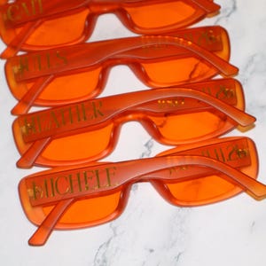 K&ouml;nnte beinhalten: Vier Paare orange get&ouml;nter Sonnenbrillen mit rechteckigen Rahmen. Jedes Paar hat einen Namen in goldfarbenen Buchstaben auf dem B&uuml;gel aufgedruckt. Die Sonnenbrillen sind leicht &uuml;berlappend angeordnet, um das Design zu zeigen.