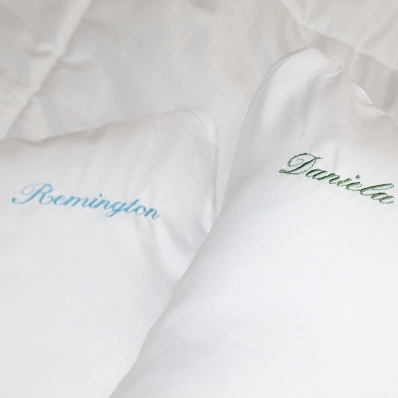 Puede incluir: Dos almohadas blancas con nombres bordados. Una almohada tiene "Daniela" bordado en verde, y la otra tiene "Remington" bordado en azul. Las almohadas est&aacute;n sobre una colcha blanca.