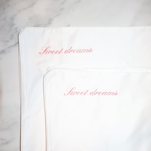 Two white pillowcases, one smaller than the other, both embroidered with "sweet dreams" in pink script.