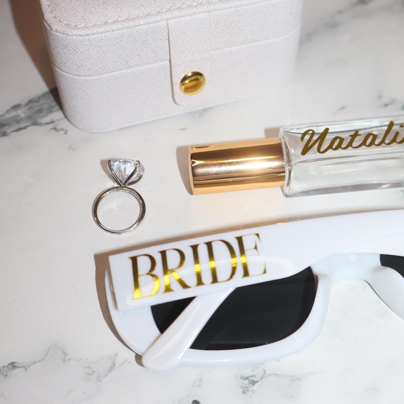K&ouml;nnte beinhalten: Eine wei&szlig;e Schmuckschatulle, Sonnenbrille mit goldfarbenem "BRIDE"-Schriftzug, ein Diamantring und eine Parf&uuml;mflasche mit goldfarbenem "Natalie"-Schriftzug. Die Artikel sind auf einer wei&szlig;en Marmoroberfl&auml;che angeordnet.