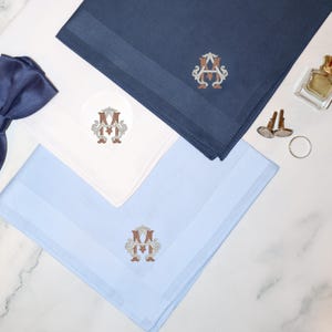 Puede incluir: Tres pañuelos de bolsillo cuadrados en azul marino, blanco y azul claro, cada uno con un monograma bordado con las letras M y A en marrón y crema. Se incluyen una pajarita azul oscuro, dos gemelos plateados y un pequeño frasco de vidrio.