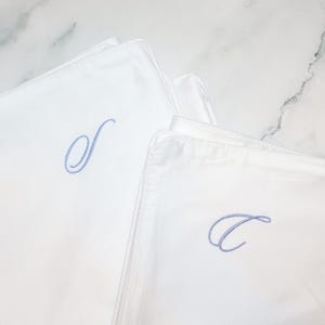 Two white pillowcases are shown, one with a light blue embroidered script "S" and the other with a light blue embroidered script "C".