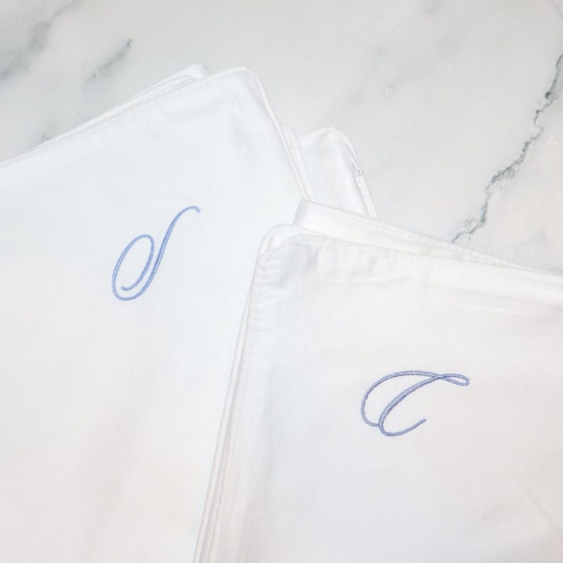 Two white pillowcases are shown, one with a light blue embroidered script "S" and the other with a light blue embroidered script "C".