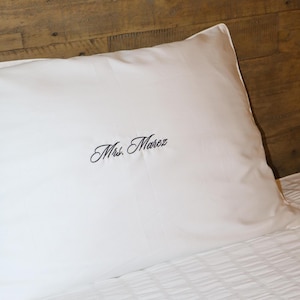A white pillowcase with "Mrs. Marez" embroidered in black script, resting on a bed with a wooden headboard.
