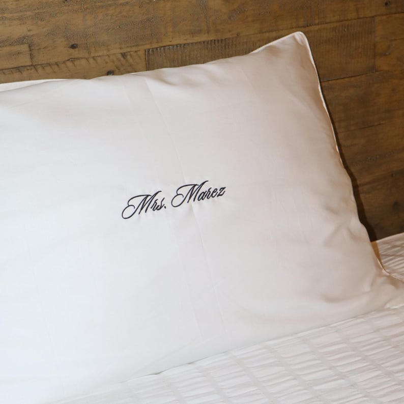 A white pillowcase with "Mrs. Marez" embroidered in black script, resting on a bed with a wooden headboard.