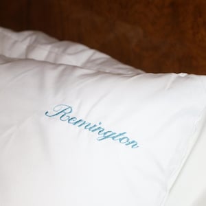 Puede incluir: Almohada blanca con el nombre "Remington" bordado en una escritura azul claro. La almohada est&aacute; hecha de una tela suave y lisa. El fondo es una superficie de madera oscura y borrosa.