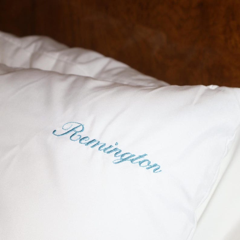 Puede incluir: Almohada blanca con el nombre "Remington" bordado en una escritura azul claro. La almohada est&aacute; hecha de una tela suave y lisa. El fondo es una superficie de madera oscura y borrosa.