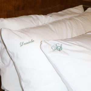 Two white pillows are on a bed with a dark wood headboard, one embroidered with "Daniela" in green script and the other with green script initials "DS".