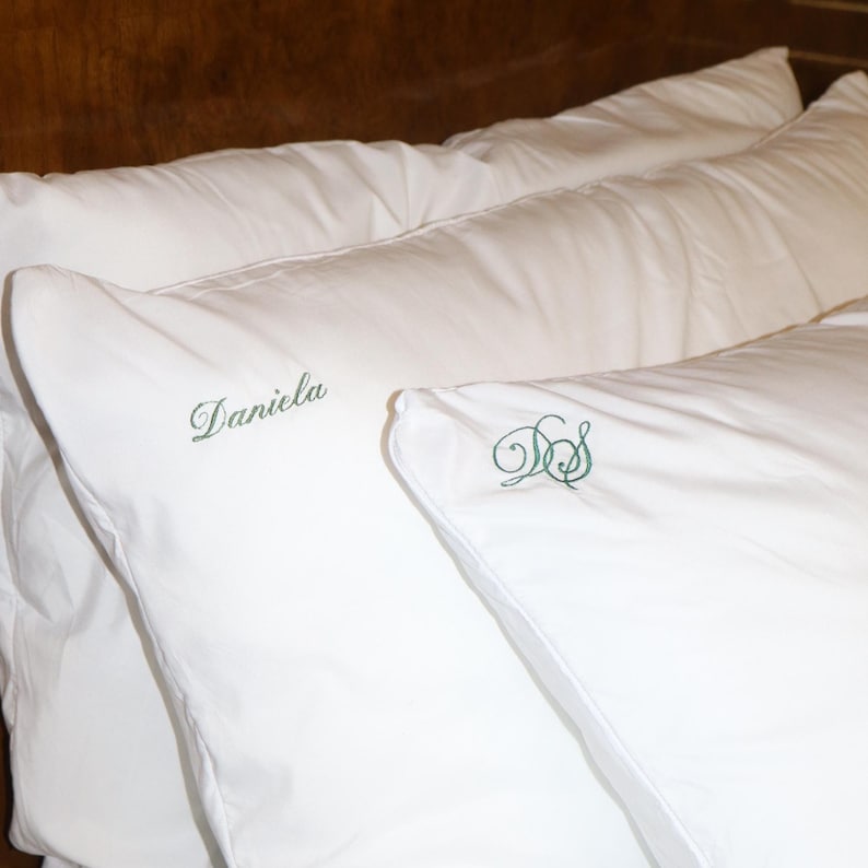 Two white pillows are on a bed with a dark wood headboard, one embroidered with "Daniela" in green script and the other with green script initials "DS".