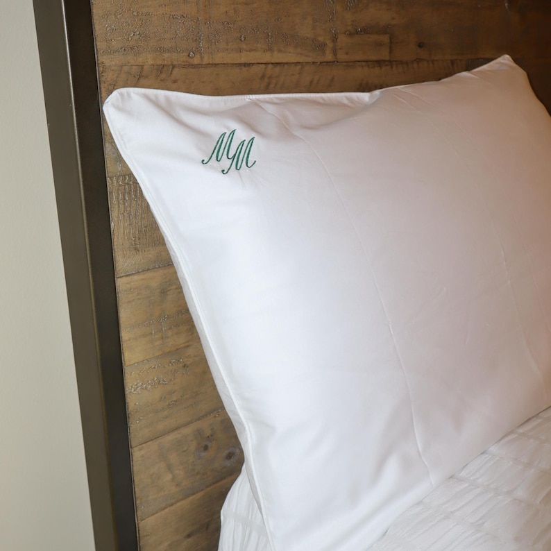 A white pillowcase with a green embroidered "M" in script, resting on a bed with a wooden headboard.