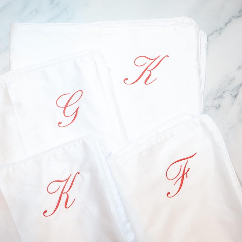 Four white pillowcases are arranged, each embroidered with a single red script initial: G, K, K, and F.