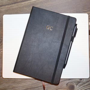 May include: A black notebook with a textured cover featuring the gold embossed letters GC, a black elastic closure band, and a black pen attached to the side, resting on top of a white rectangular notepad with a brown ribbon bookmark visible.