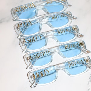 K&ouml;nnte beinhalten: Brillen mit transparentem Rahmen und hellblauen Gl&auml;sern. Jede Brille hat einen Namen in goldfarbenen Buchstaben auf dem Rahmen. Die Namen sind Shawna, Julia, Ashlly, Charlotte und Molly. Die Brillen sind &uuml;bereinander gestapelt.