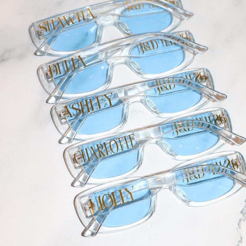 K&ouml;nnte beinhalten: Brillen mit transparentem Rahmen und hellblauen Gl&auml;sern. Jede Brille hat einen Namen in goldfarbenen Buchstaben auf dem Rahmen. Die Namen sind Shawna, Julia, Ashlly, Charlotte und Molly. Die Brillen sind &uuml;bereinander gestapelt.