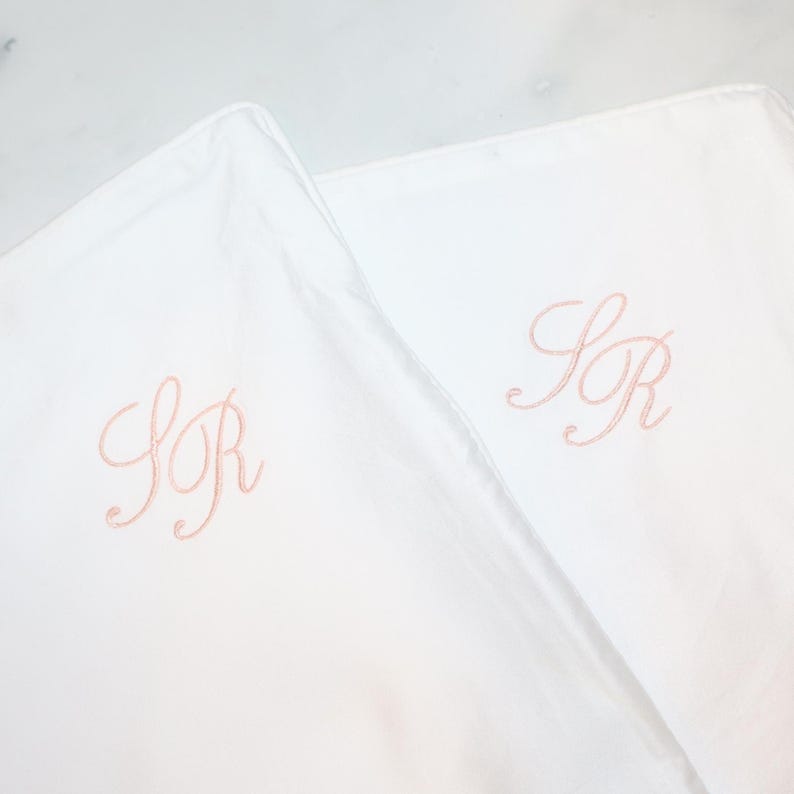 Puede incluir: Dos fundas de almohada blancas con iniciales bordadas en un color rosa suave. Las iniciales "SR" se muestran elegantemente en cada funda de almohada, ofreciendo un toque personalizado. La ropa de cama tiene una apariencia suave y n&iacute;tida.
