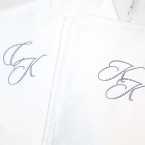 Puede incluir: Dos fundas de almohada blancas con las iniciales "JK" bordadas en gris claro. Las iniciales est&aacute;n en una fuente cursiva. Las fundas de almohada est&aacute;n dobladas y dispuestas una al lado de la otra.