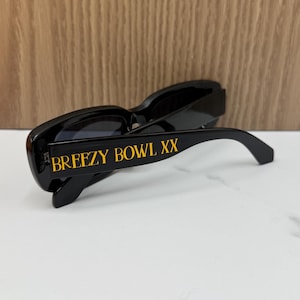 K&ouml;nnte beinhalten: Schwarze Sonnenbrille mit goldfarbenem Schriftzug "BREEZY BOWL XX" auf dem B&uuml;gel. Die Sonnenbrille hat einen rechteckigen Rahmen und dunkle Gl&auml;ser. Das Bild ist aus einem niedrigen Winkel aufgenommen.