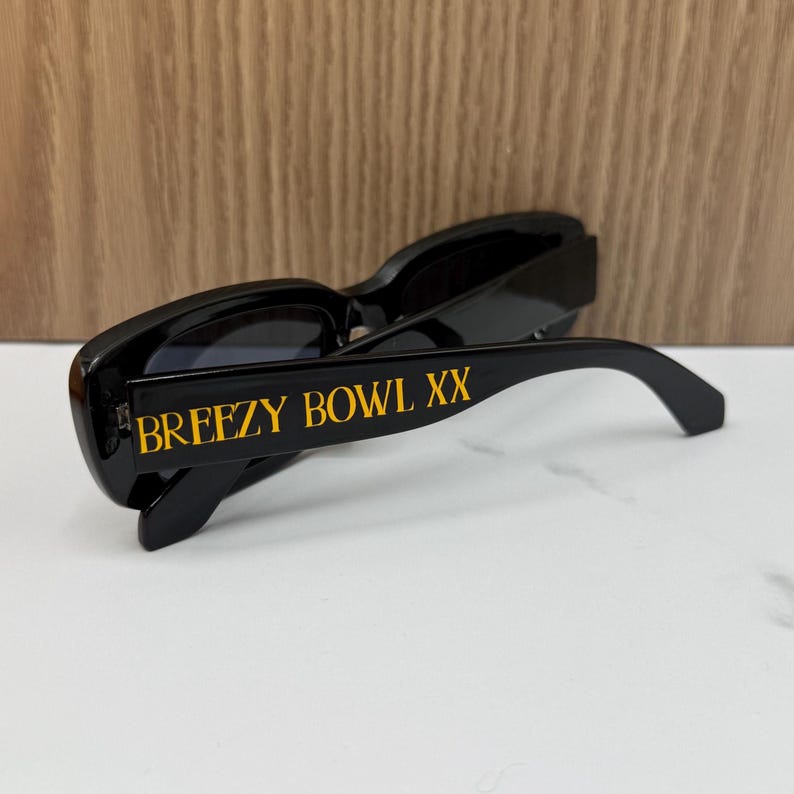 K&ouml;nnte beinhalten: Schwarze Sonnenbrille mit goldfarbenem Schriftzug "BREEZY BOWL XX" auf dem B&uuml;gel. Die Sonnenbrille hat einen rechteckigen Rahmen und dunkle Gl&auml;ser. Das Bild ist aus einem niedrigen Winkel aufgenommen.
