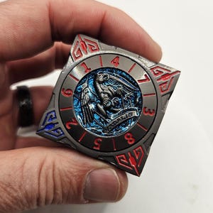 Może przedstawiać: Kwadratowa, metalowa moneta o szczeg&oacute;łowym wzorze. Moneta posiada centralny niebieski emblemat z ptakiem i napisem "VADALIS". Zewnętrzny pierścień ma cyfry oraz czerwone i niebieskie akcenty.
