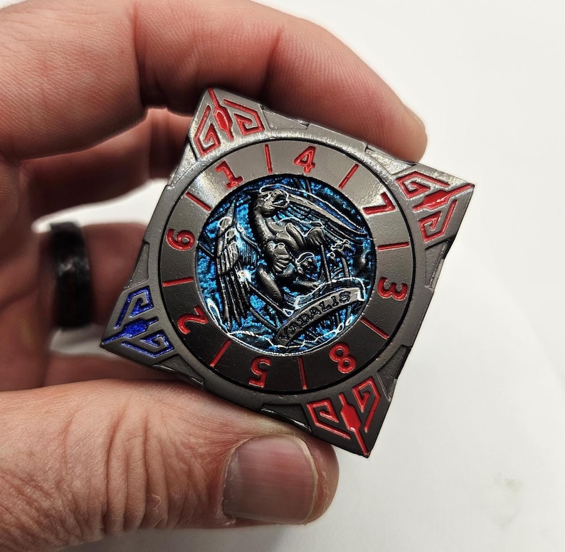 Może przedstawiać: Kwadratowa, metalowa moneta o szczeg&oacute;łowym wzorze. Moneta posiada centralny niebieski emblemat z ptakiem i napisem "VADALIS". Zewnętrzny pierścień ma cyfry oraz czerwone i niebieskie akcenty.
