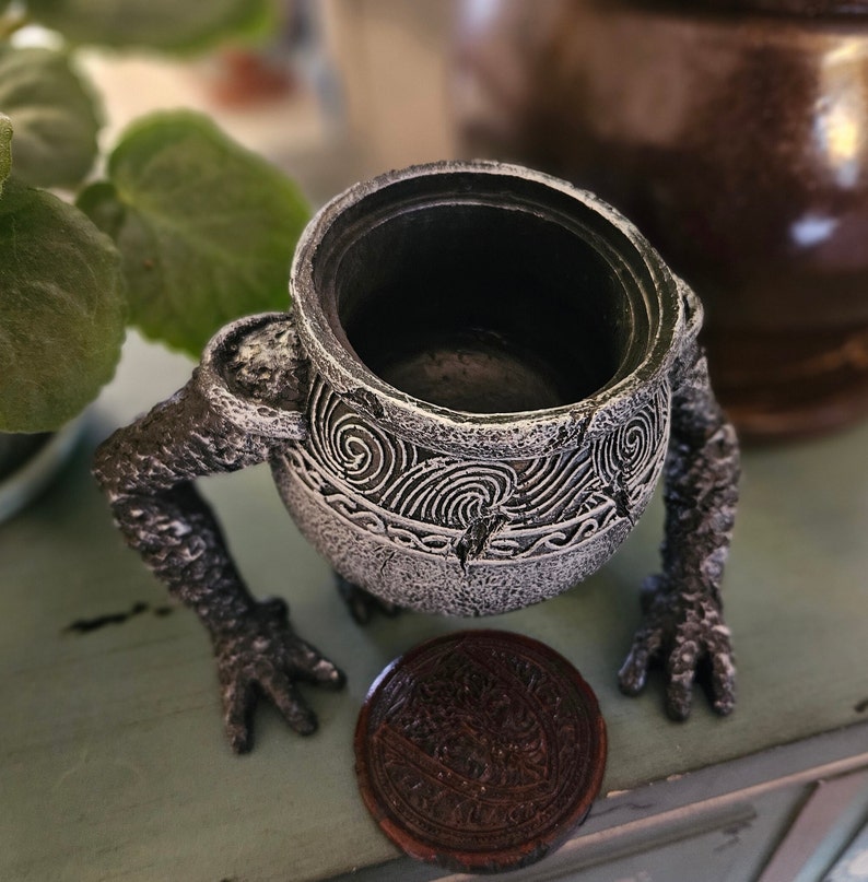 May include: A small, gray, ceramic cauldron with a textured surface and intricate designs. The cauldron has two arms and is sitting on a wooden surface.