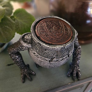 May include: A gray ceramic frog figurine with a detailed design. The frog is holding a small, round, copper-colored dish with a decorative pattern.