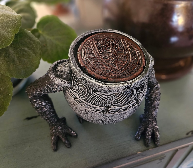 May include: A gray ceramic frog figurine with a detailed design. The frog is holding a small, round, copper-colored dish with a decorative pattern.