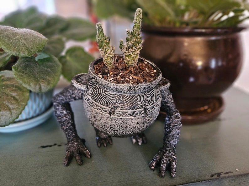 May include: A gray ceramic planter shaped like a creature with two arms and legs. The planter has a textured surface and is decorated with a swirling pattern. A small cactus plant is growing in the planter.