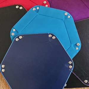 May include: A collection of hexagonal, foldable dice trays in various colors, including blue, navy, red, black, and purple. Each tray is constructed with a stitched border and metal snap closures at the corners. The trays are designed for tabletop gaming.