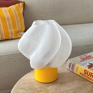 May include: A table lamp featuring a white, spiraled, ribbed shade with a cylindrical yellow base. The lamp has a distinct twisted, organic shape and sits on a round surface next to a book titled Surf Photography with a red and yellow cover.