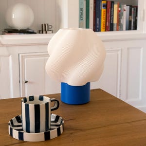May include: A white 3D-printed table lamp with a spiraled, wavy shade and a solid blue cylindrical base, positioned next to a white ceramic cup and saucer featuring thick, vertical navy blue stripes on a wooden surface.
