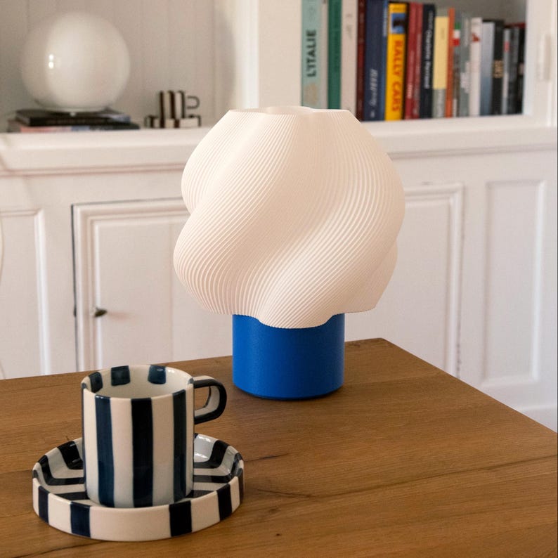 May include: A white 3D-printed table lamp with a spiraled, wavy shade and a solid blue cylindrical base, positioned next to a white ceramic cup and saucer featuring thick, vertical navy blue stripes on a wooden surface.