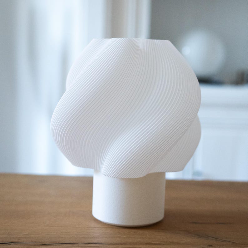 May include: A white vase with a textured, spiraling, ribbed upper section and a smooth, cylindrical base. The object features a distinct, twisted geometric form that creates a series of diagonal lines across its surface, standing upright.